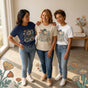 Three diverse mothers wearing matching Matrescence shirts in different colors for Mother's Day 2026.