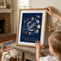 A mother adjusting the gold frame of the Mama Est 2026 print as her child looks on
