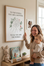 A high-resolution professional photo print in a light oak framed matted poster on a warm nursery wall, displaying the exact quote text 'To the World, You Are a Mother. To Us, You Are the World.' above a minimalist family hands line art holding hands surrounded by a watercolor eucalyptus and pink floral wreath. A joyful mother with wavy brown hair in a cozy knitted sweater holds her small baby in her arms, looking at the poster with heartwarming happiness. Her right hand gently touches the frame. The low she